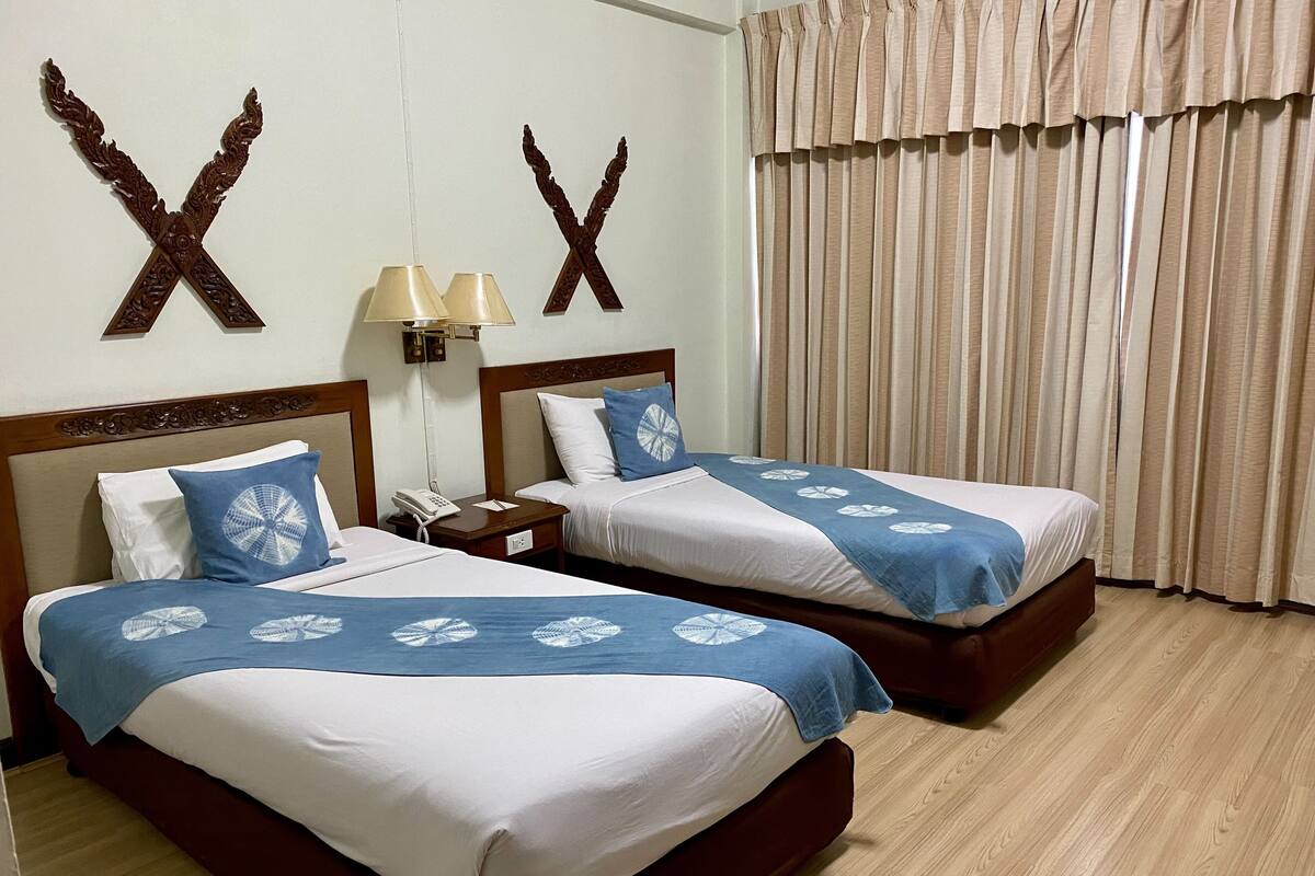 Habitación de hotel con dos camas, cada una con cubrecama azul y blanco, cabecero de madera, mesita de noche con lámpara y ventana con cortinas color beige.