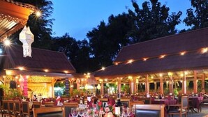 Un área de comedor al aire libre con mesas y sillas de madera, luces colgantes y una zona de estar cubierta.