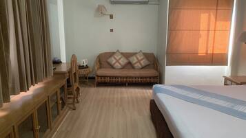A hotel room with a wooden desk, a sofa with patterned cushions, a bed, and a window with blinds.