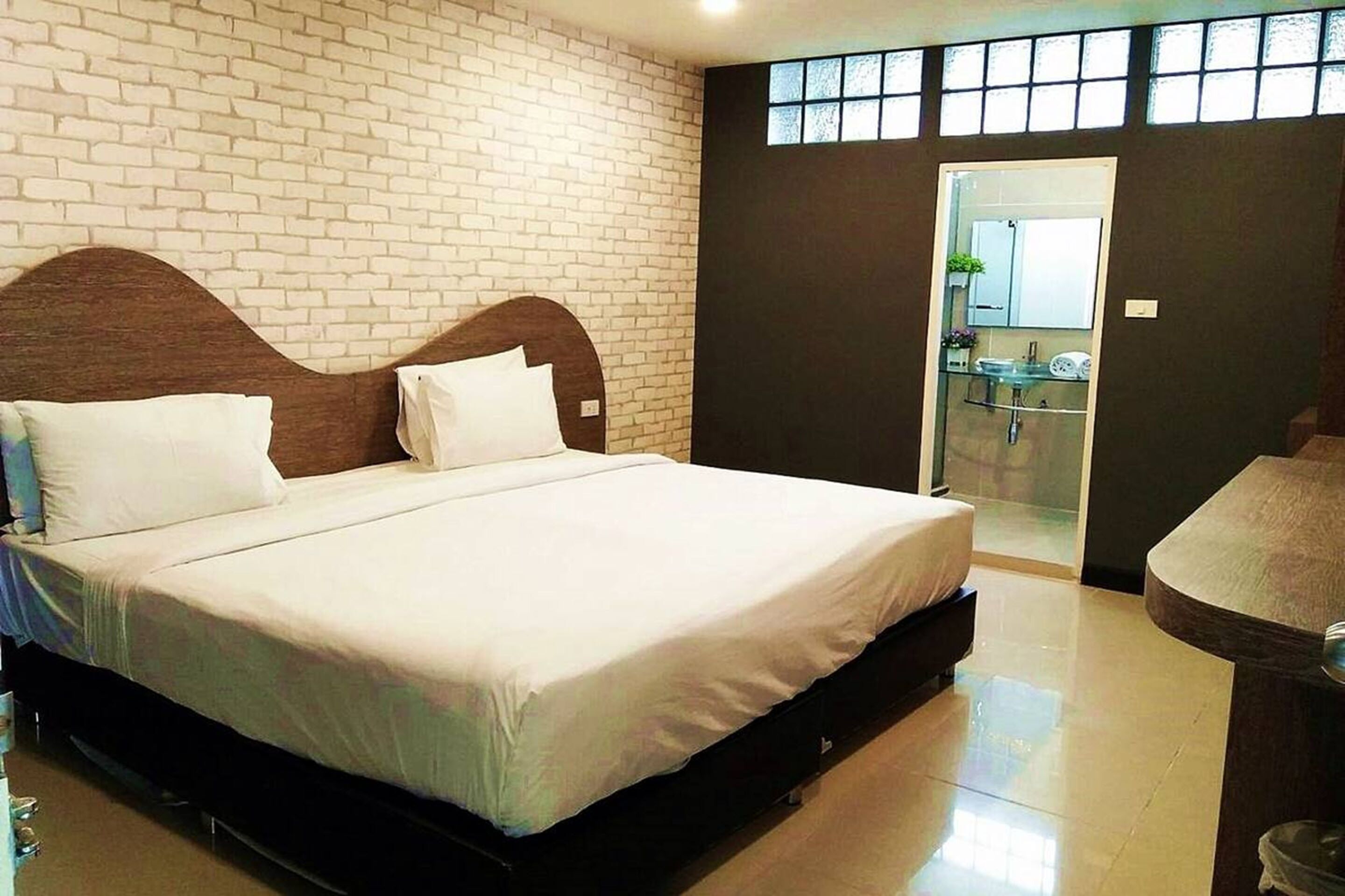 A hotel room with a large bed, a wooden headboard, a bathroom with a glass shower, and a view of the outside through a window.