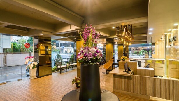 Um hall de hotel moderno com piso de madeira, balcão de recepção e um vaso preto grande com flores.