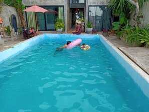 Una piscina con dos niños en el agua, rodeada de plantas en macetas y mobiliario de exterior.