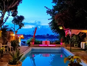 Zona de piscina con vista a un cuerpo de agua, rodeada de plantas en macetas y mobiliario de exterior.