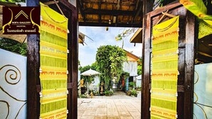 Une porte d’entrée ornée de deux panneaux de tissu jaune à motifs, de part et d’autre, qui donne sur une cour dallée et verdoyante.