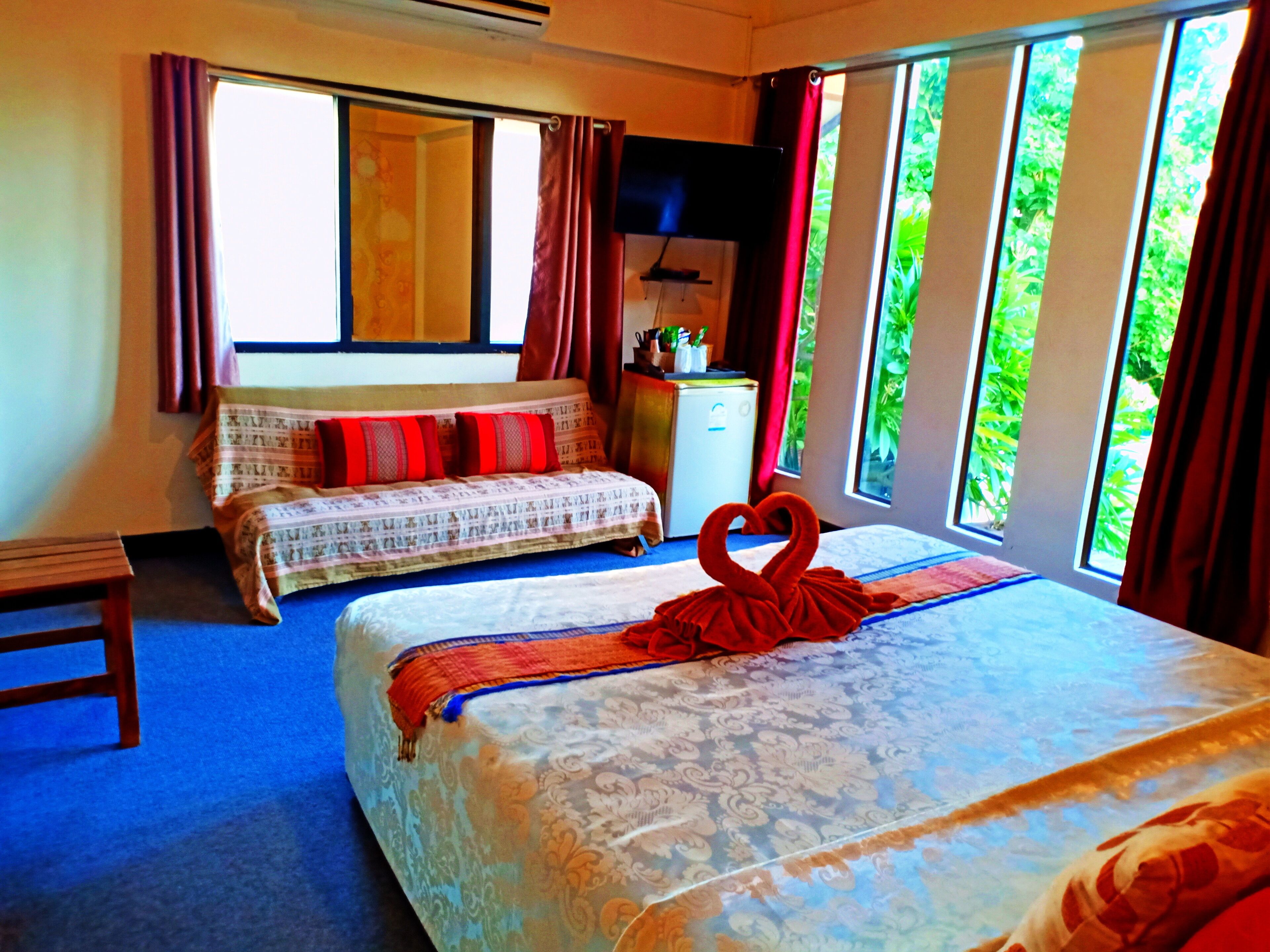A hotel room with two beds, a desk, a window with curtains, and a view of greenery outside.