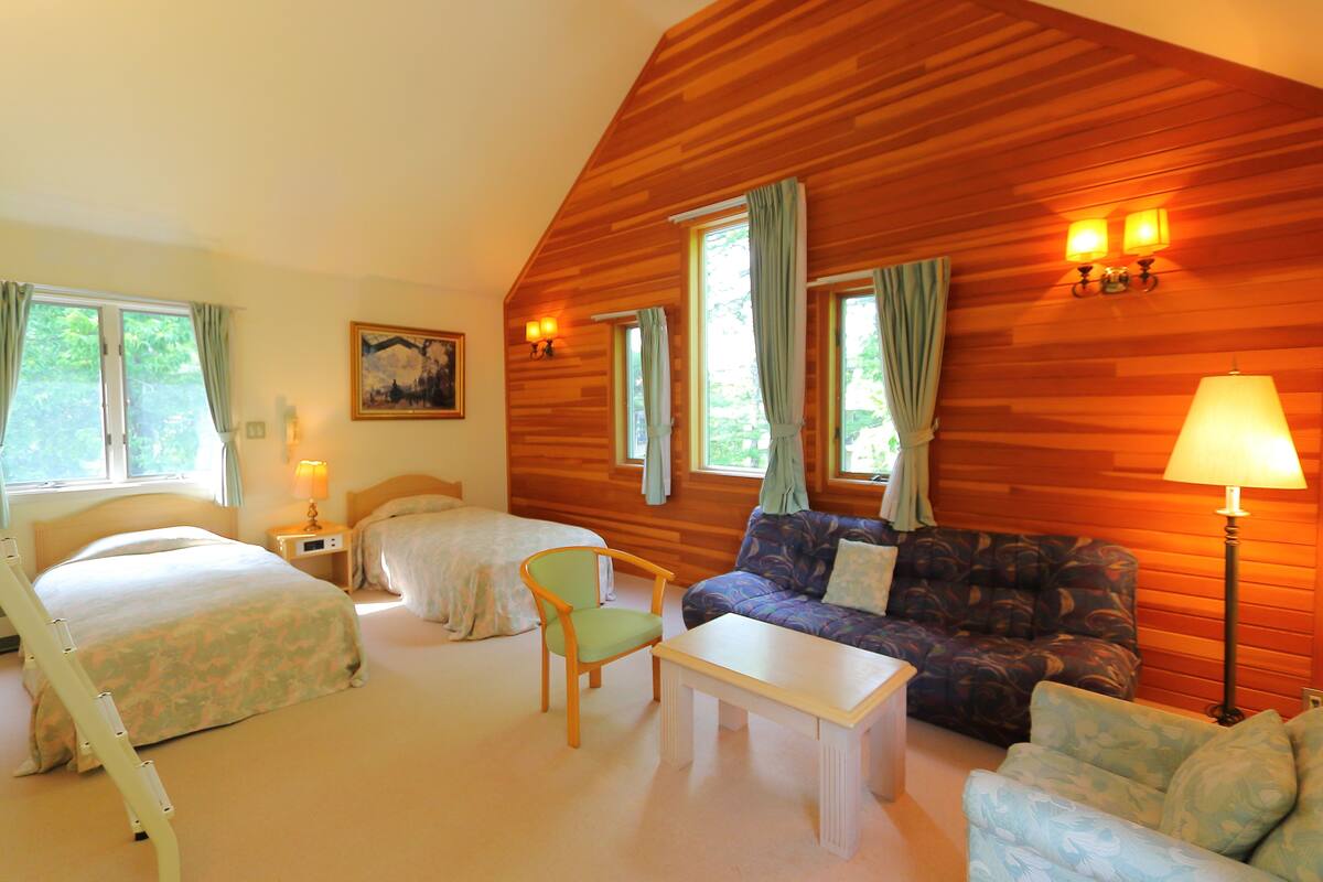 A room with two beds, a sofa, a chair, and a table. There are windows with curtains, a painting on the wall, and wall-mounted lamps.