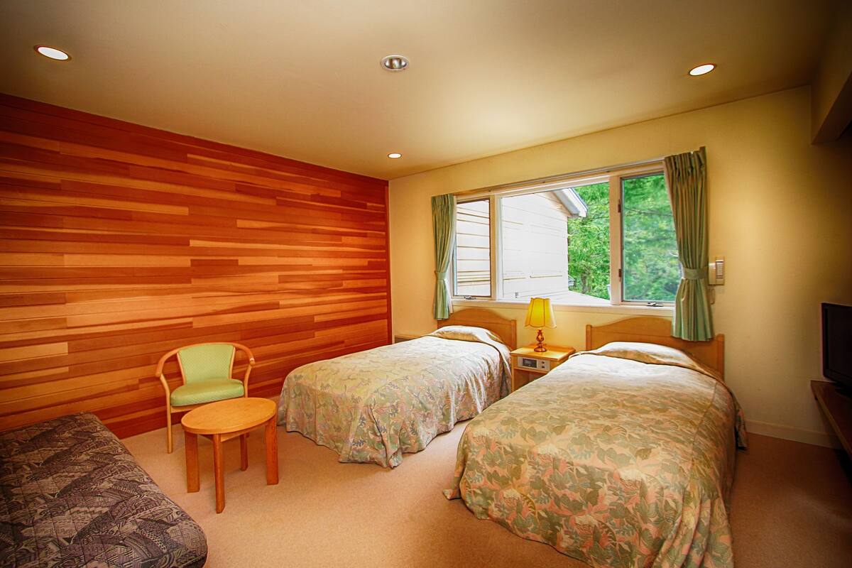 A hotel room with two beds, a wooden wall, a window with curtains, a chair, a small table, and a TV.
