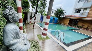 A serene outdoor pool area with a Buddha statue, striped poles, and a modern building in the background.