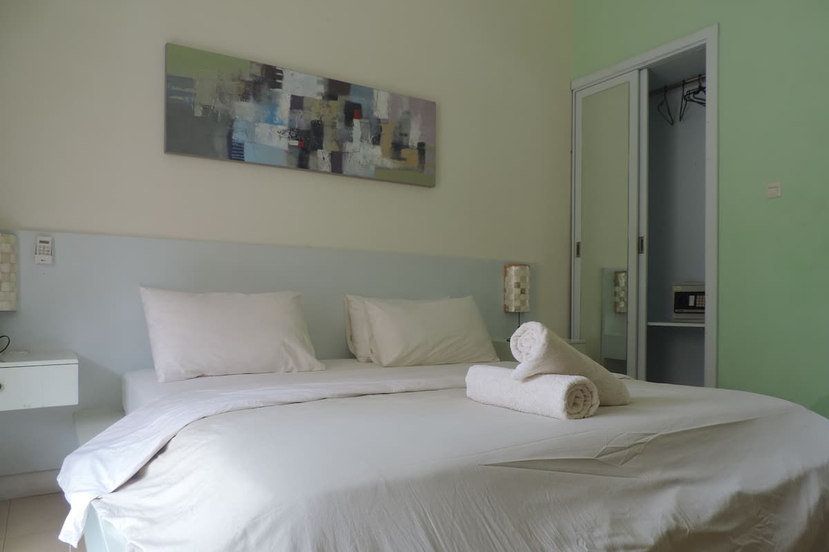 A neatly made bed with white linens and a rolled towel, a bedside table with a lamp, and a painting on the wall.