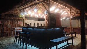 Un bar frente a la playa con techo de paja, taburetes de madera y una barra.