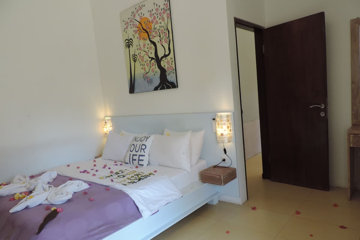 A neatly made bed with a white and purple bedspread, a pillow with "ENJOY YOUR LIFE" text, and a wall-mounted painting of a tree.