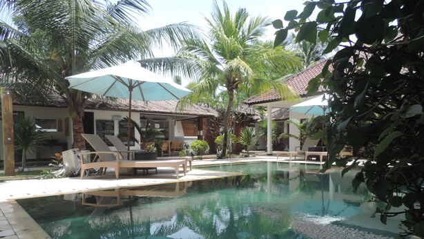 Zona de piscina con sillones de descanso y sombrillas, rodeada de palmeras y una estructura de techo con tejas.
