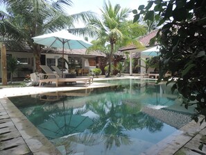 A pool area with lounge chairs and umbrellas, surrounded by palm trees and a tiled roof structure. - Casus Dream Hotel (Gili Trawangan)