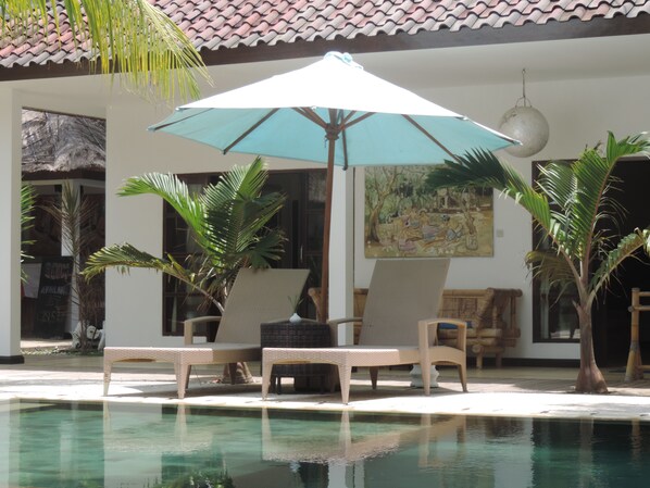 Un área junto a la piscina con sillones de descanso y una sombrilla grande, rodeada de palmeras y un techo de tejas.