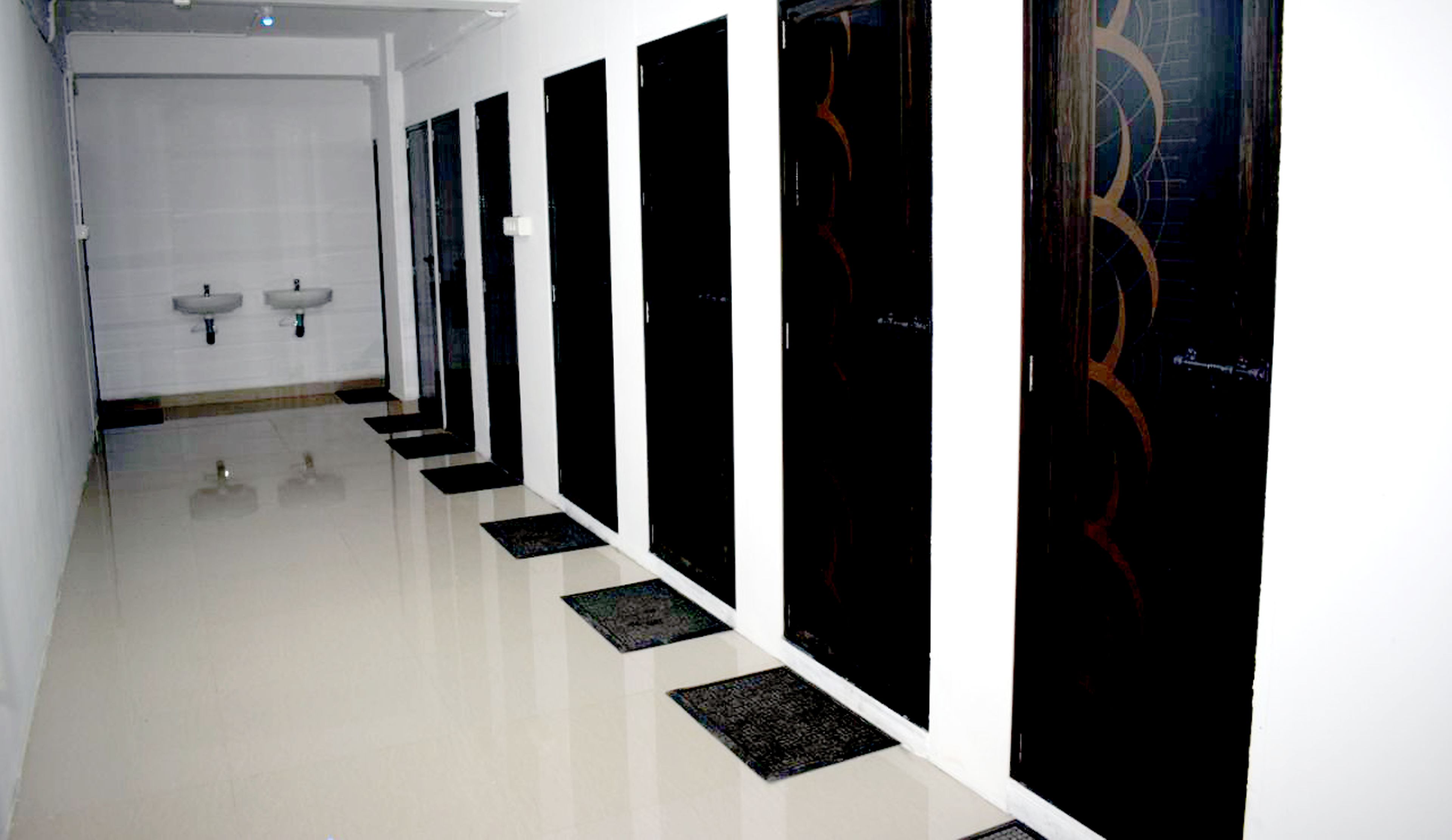 A long, narrow hallway with black doors and white walls. There are two sinks visible at the end of the hallway.