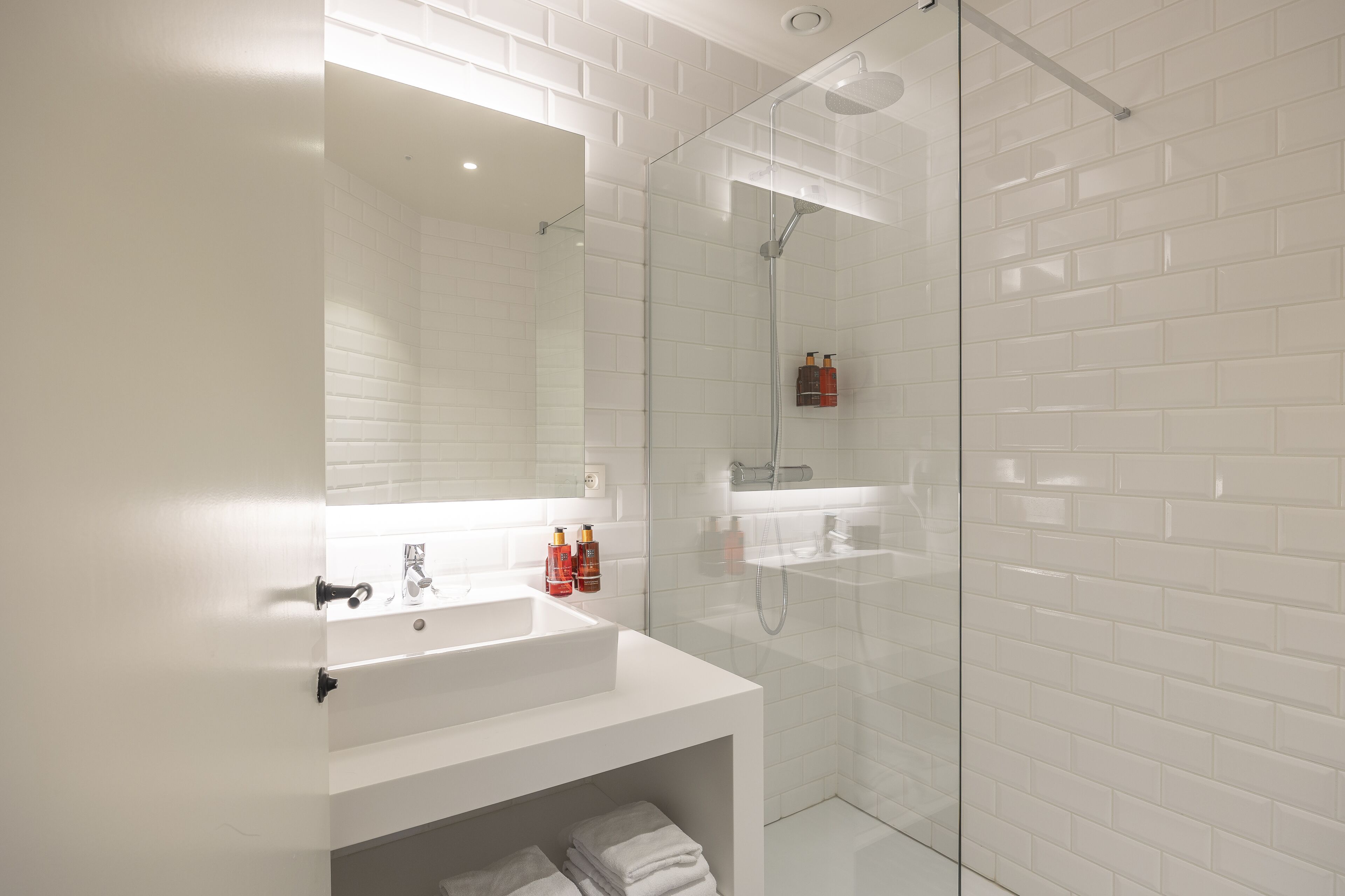 Un baño moderno con lavamanos blanco, cabina de ducha de vidrio y paredes con azulejos blancos.