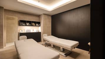 A clinical examination room with two examination tables, a desk with medical supplies, and a shelf with personal items.