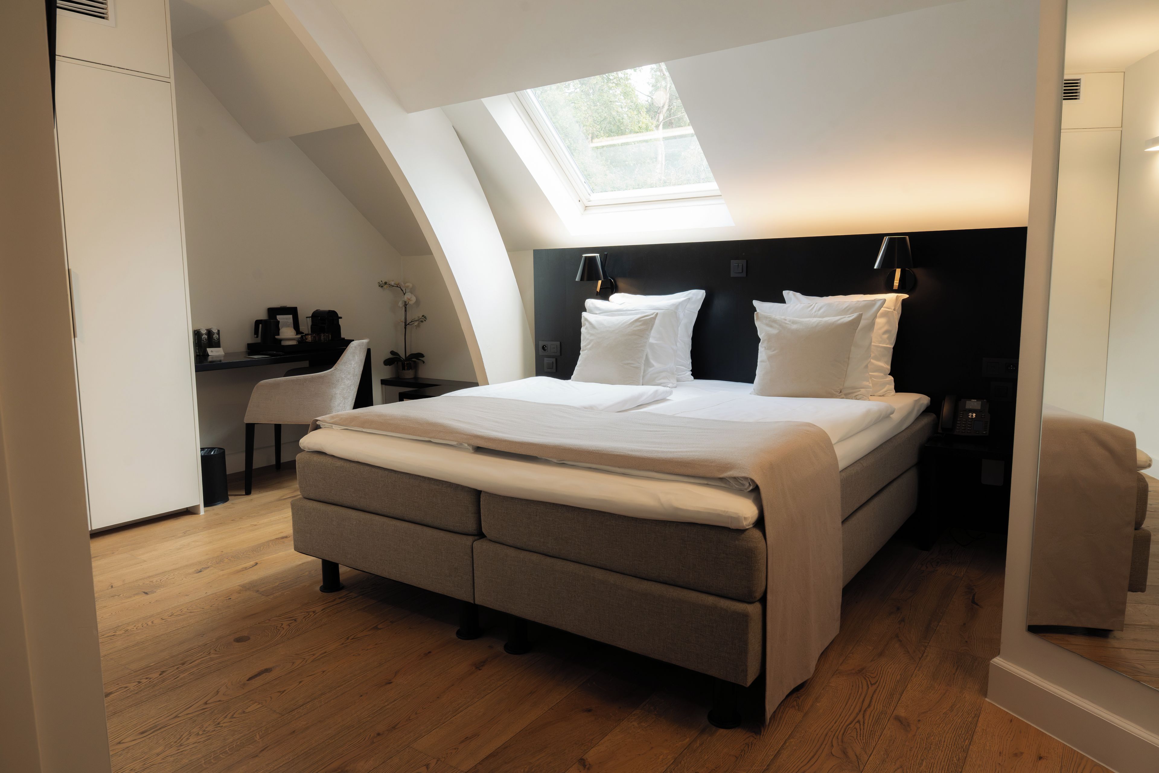 Une chambre avec un puits de lumière, un lit avec du linge de maison blanc, un bureau avec une chaise et une petite plante en pot.