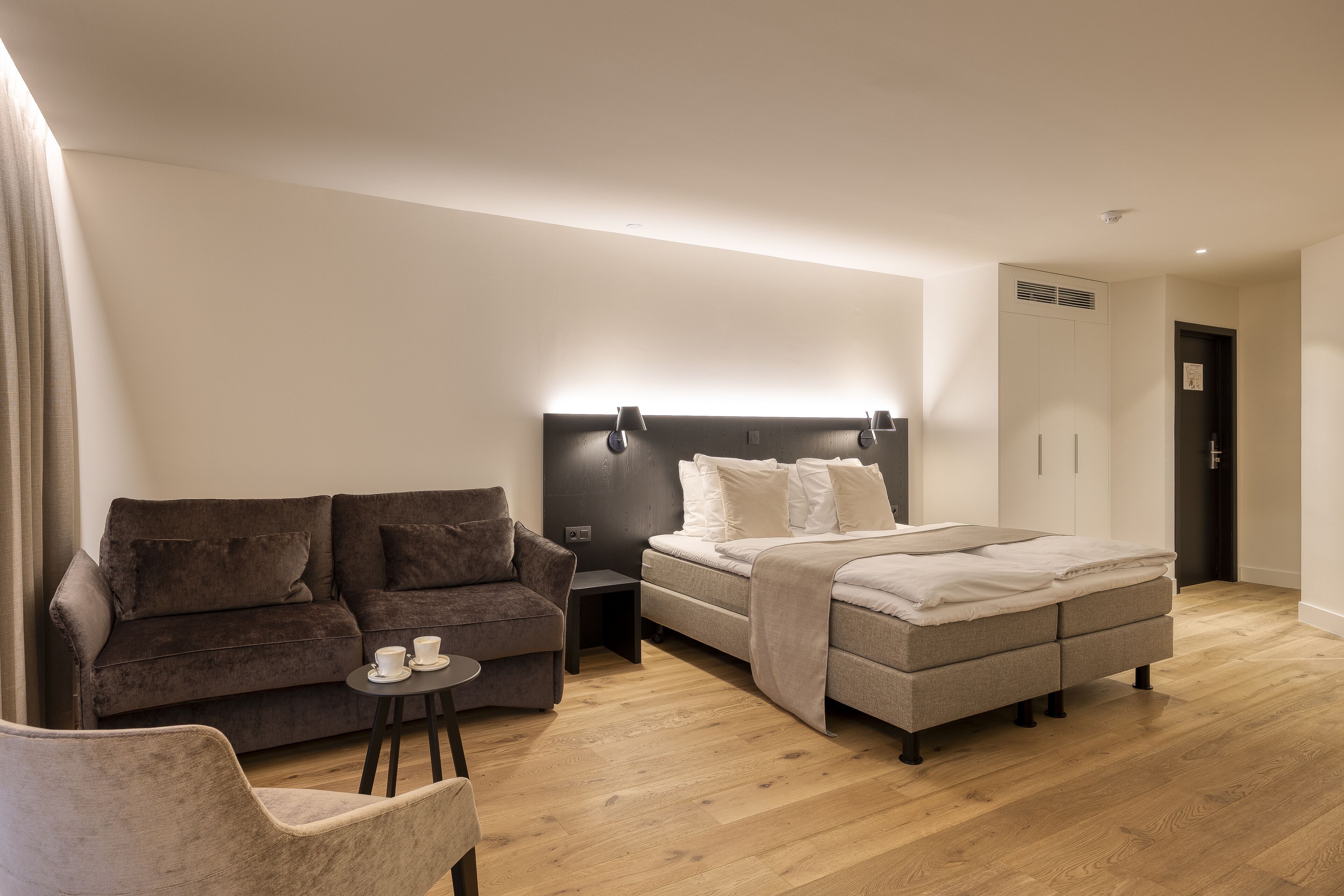 A modern hotel room with a sofa, a small table with a cup, a bed with a headboard, and a wardrobe.