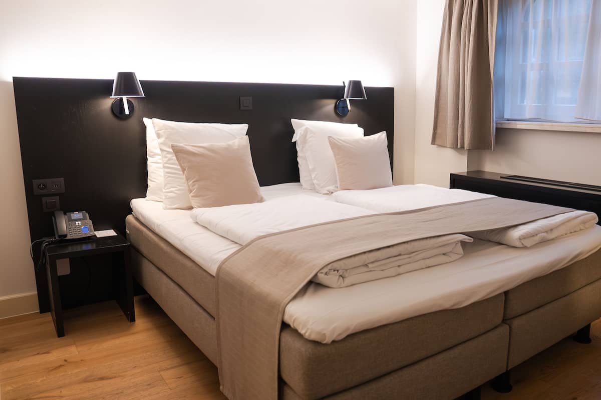 Una cama bien hecha con cabecera, dos lámparas de noche, un teléfono en la mesita y una ventana con cortinas.