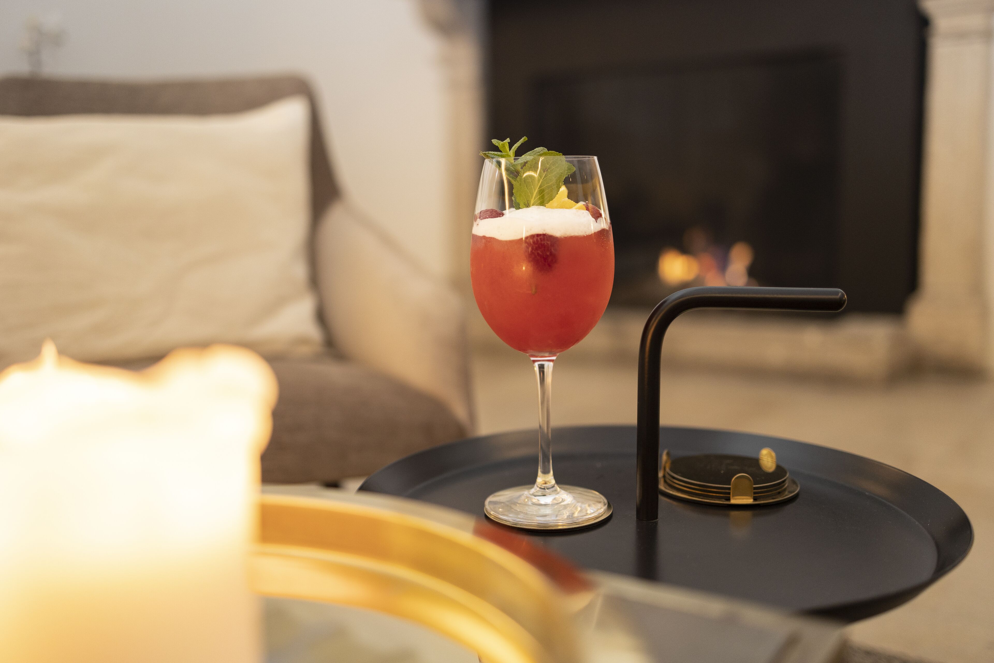 A glass of red beverage with garnish on a black table, a lit candle, and a fireplace in the background.