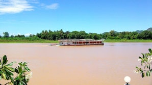 A riverboat on a calm river with lush greenery and hills in the background. - Khunya Resort Riverkwai (Kanchanaburi)
