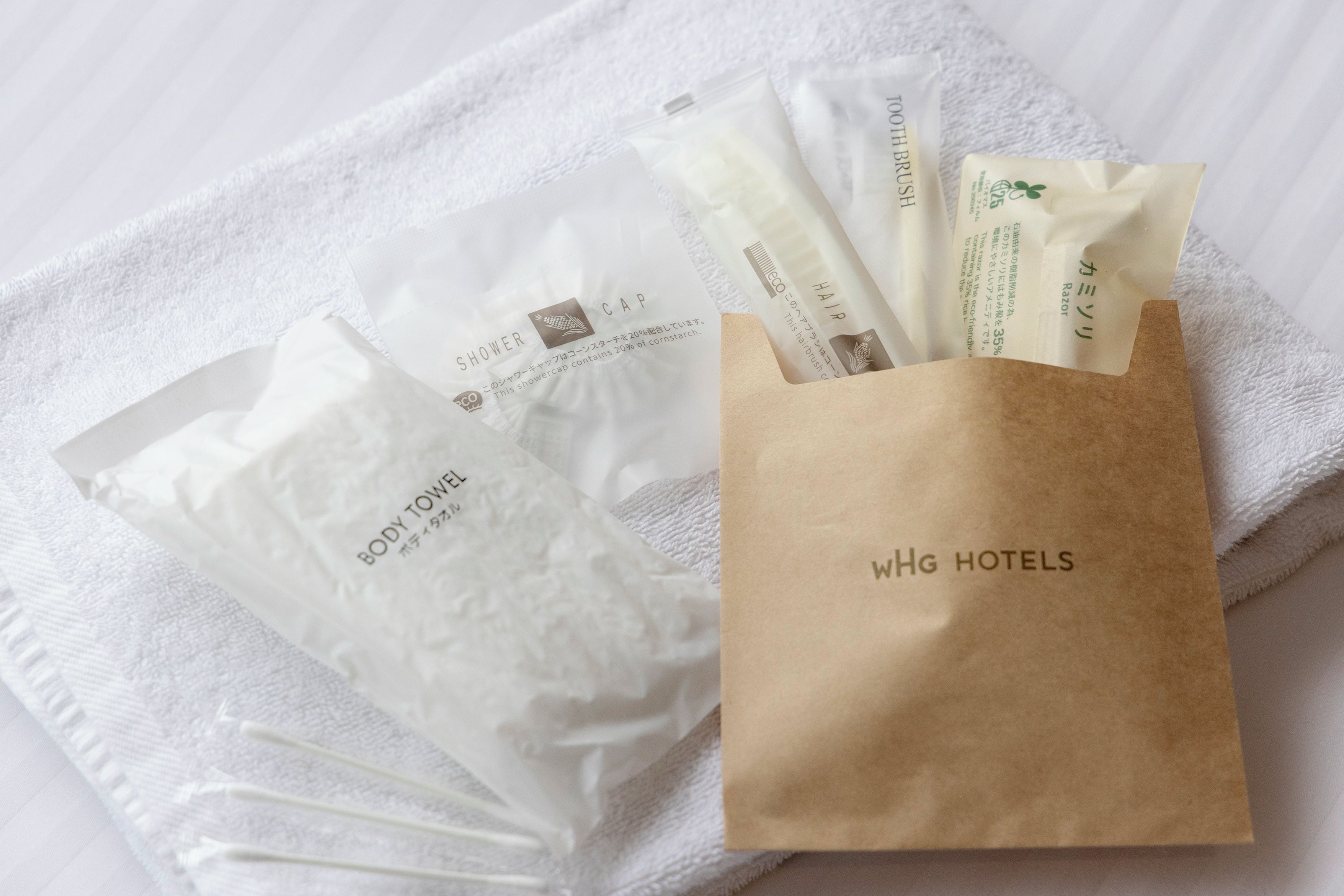 A set of toiletries including a body towel, a shower cap, and a hairbrush, all packaged in a brown paper bag with 'WHG HOTELS' branding.