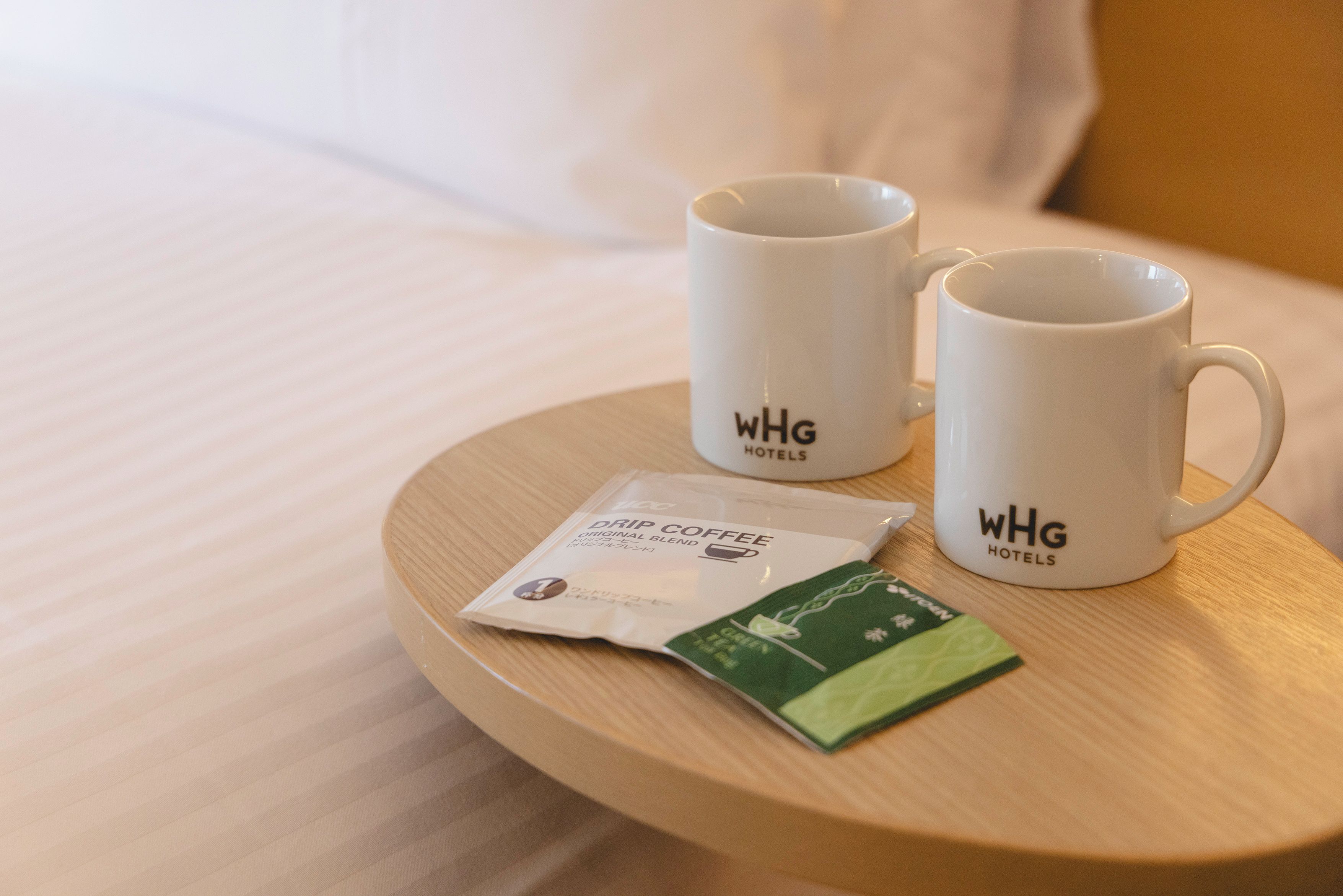 Duas canecas brancas com a marca WHG Hotels sobre uma bandeja de madeira, juntamente com um sachê de café filtrado e um sachê de chá verde.