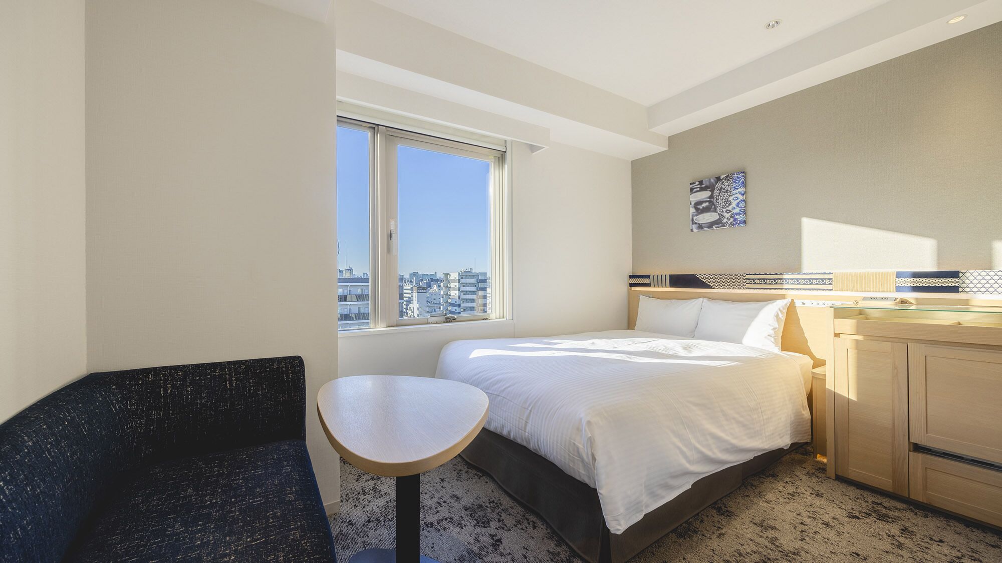 A hotel room with a bed, a small table, a chair, a window with a city view, and a bedside table with a lamp.