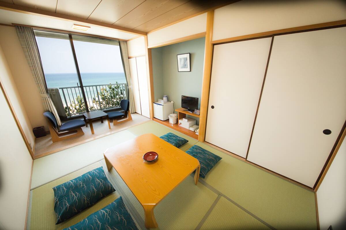 Ein Zimmer mit Tatami-Boden, einem niedrigen Holztisch und Kissen. Eine Schiebetür führt auf einen Balkon mit Meerblick.
