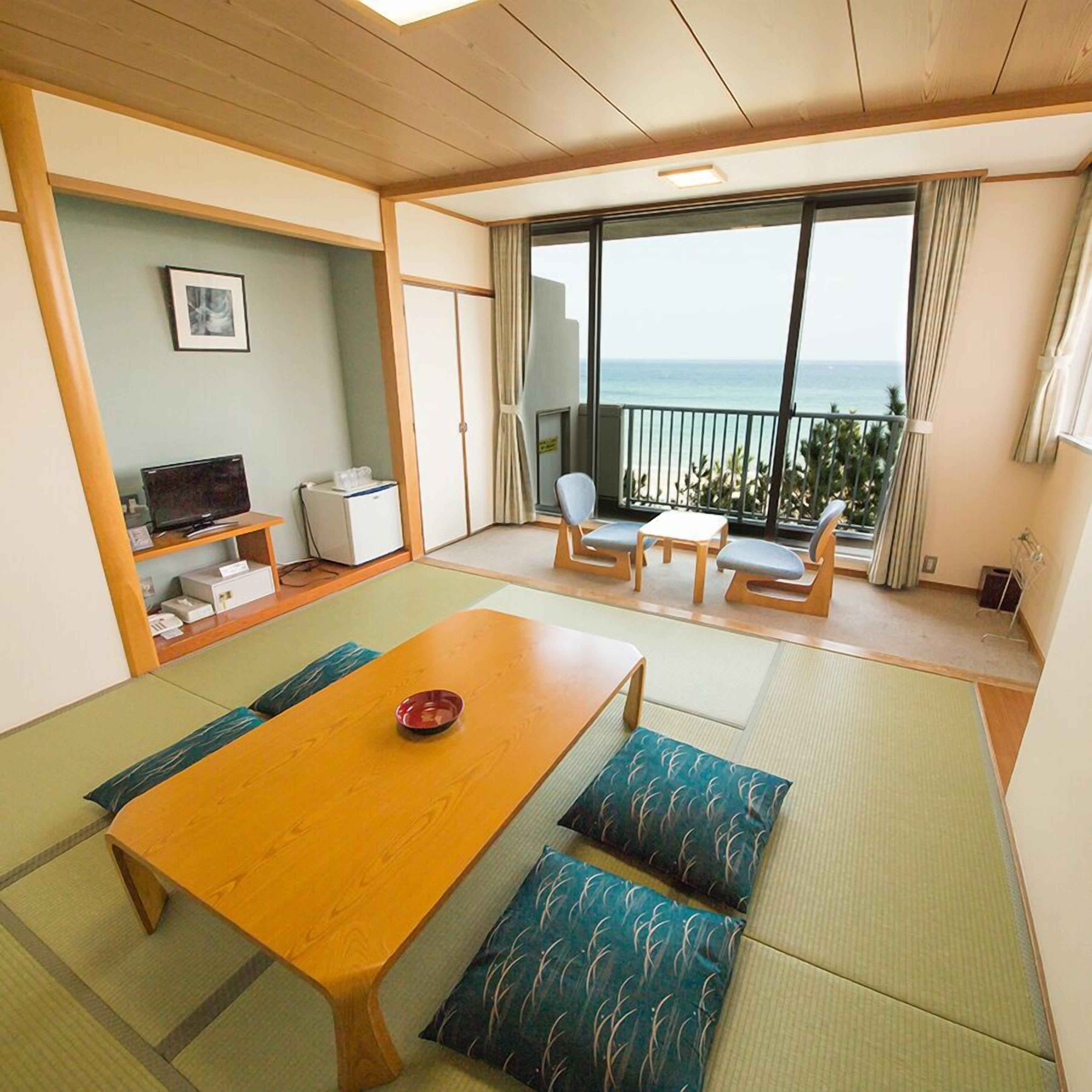 A room with a wooden table, cushioned seating, a TV, a small fridge, and a balcony with a view of the sea.
