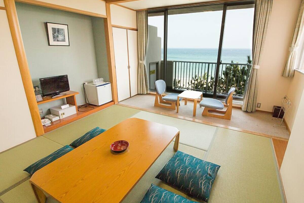 Ein Zimmer mit Holztisch, gepolsterten Sitzgelegenheiten, Fernseher, kleinem Kühlschrank und einem Balkon mit Meerblick.