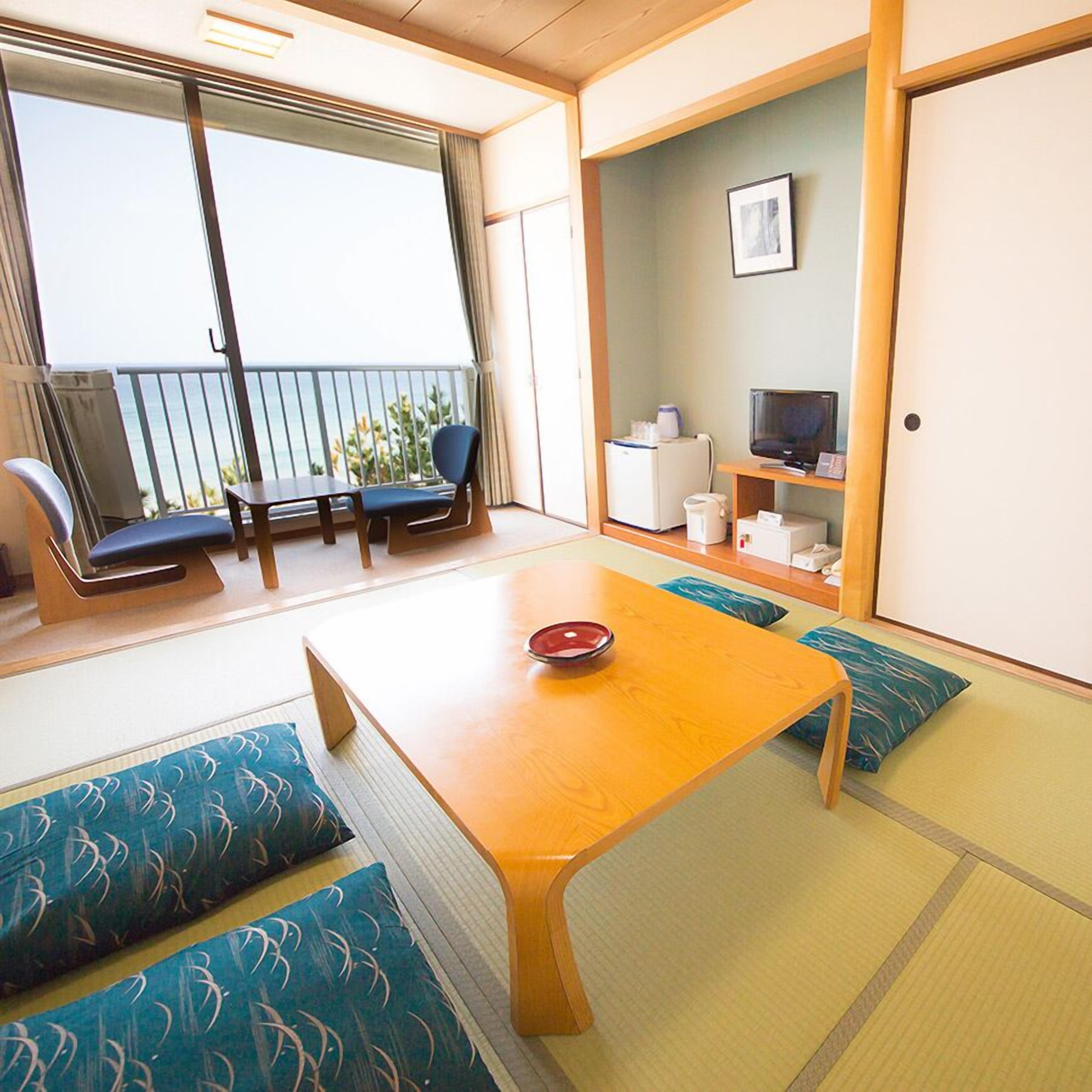 A room with tatami flooring, a wooden table with a red bowl, a TV, a refrigerator, and a balcony with a view of the ocean.