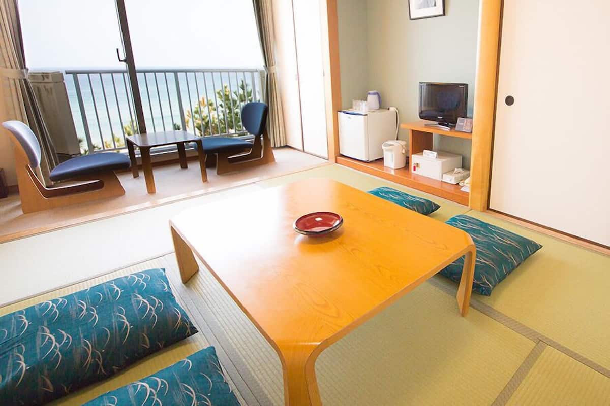 Ein Zimmer mit Tatami-Boden, einem Holztisch mit roter Schüssel, einem Fernseher, einem Kühlschrank und einem Balkon mit Meerblick.