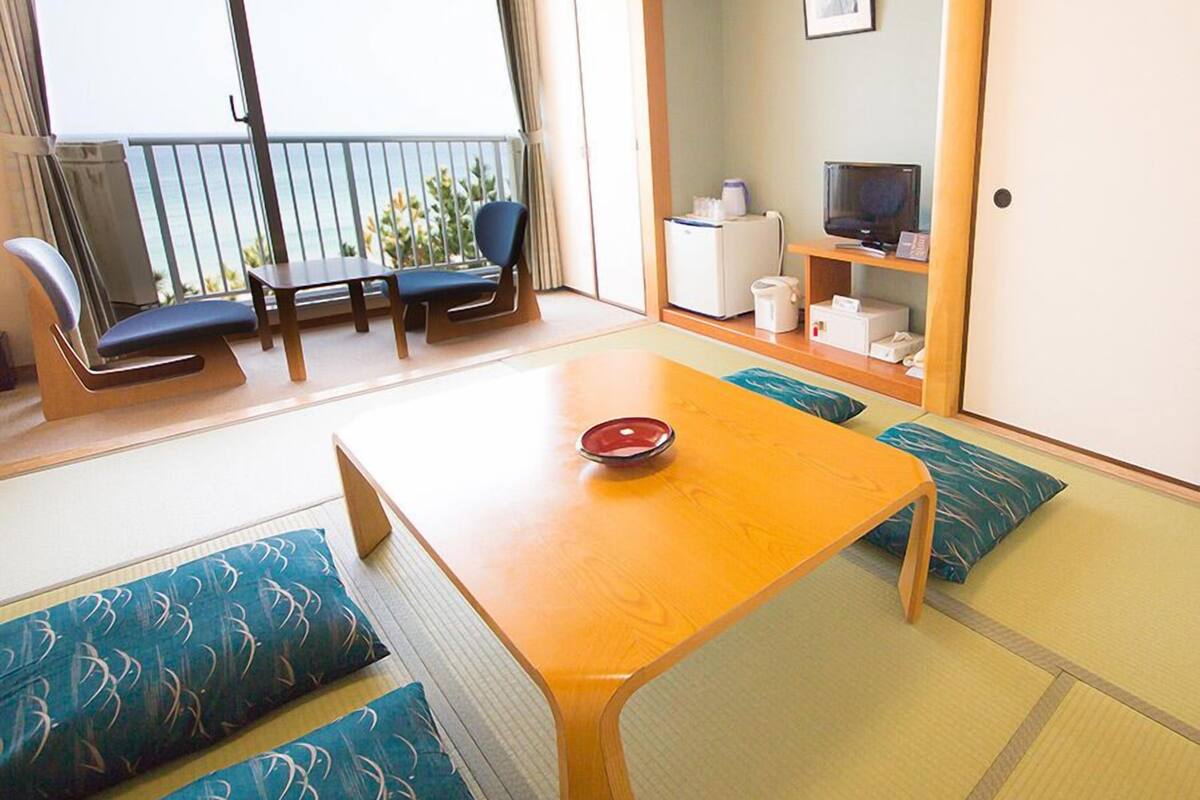 Ein Zimmer mit Tatami-Boden, einem Holztisch mit roter Schüssel, einem Fernseher, einem Kühlschrank und einem Balkon mit Meerblick.