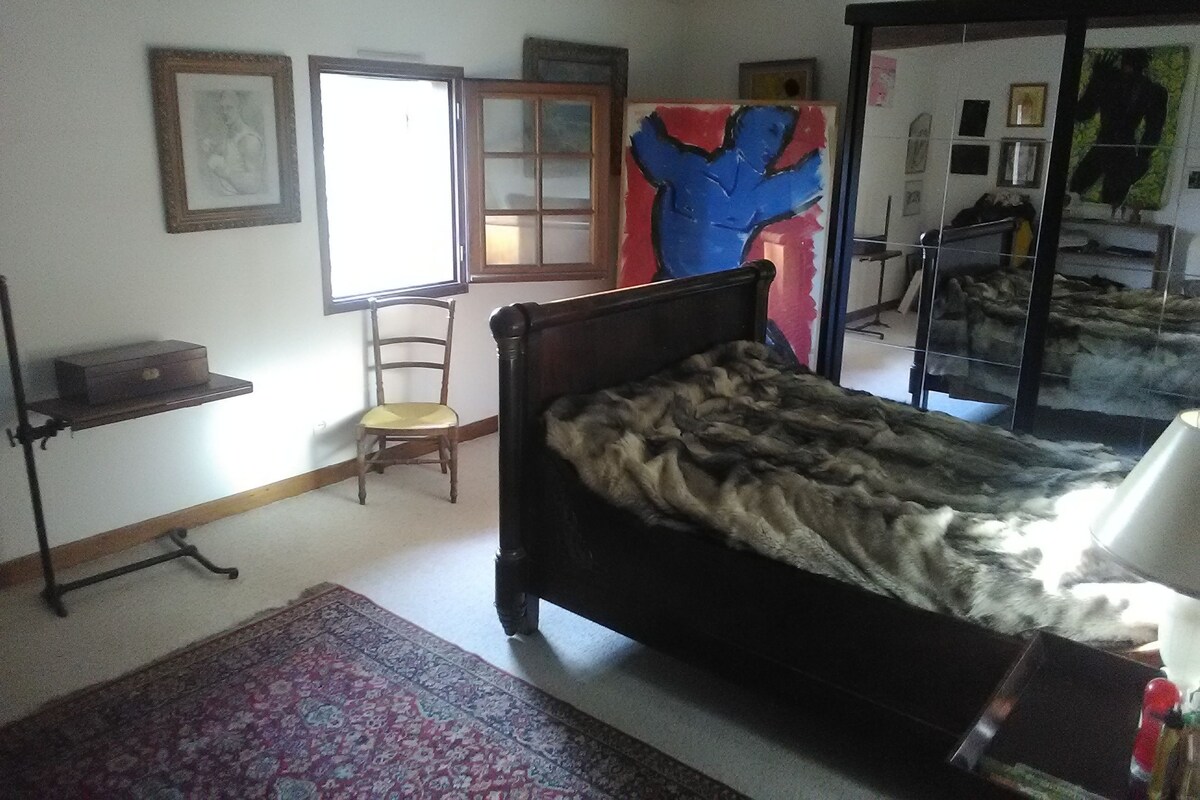 A bedroom with a bed, a chair, a shelf, a window, and a mirror.