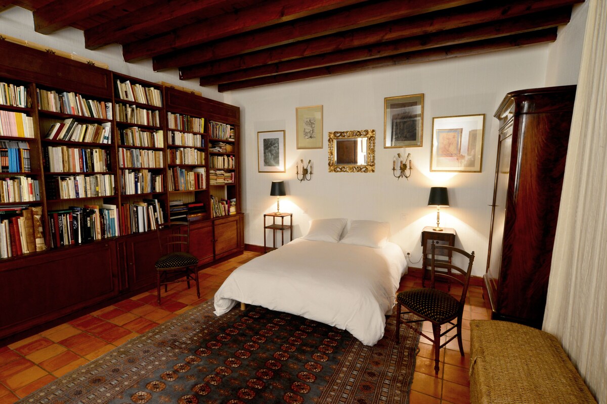 A room with a large bookshelf, two bedside tables with lamps, a bed, and a patterned rug.