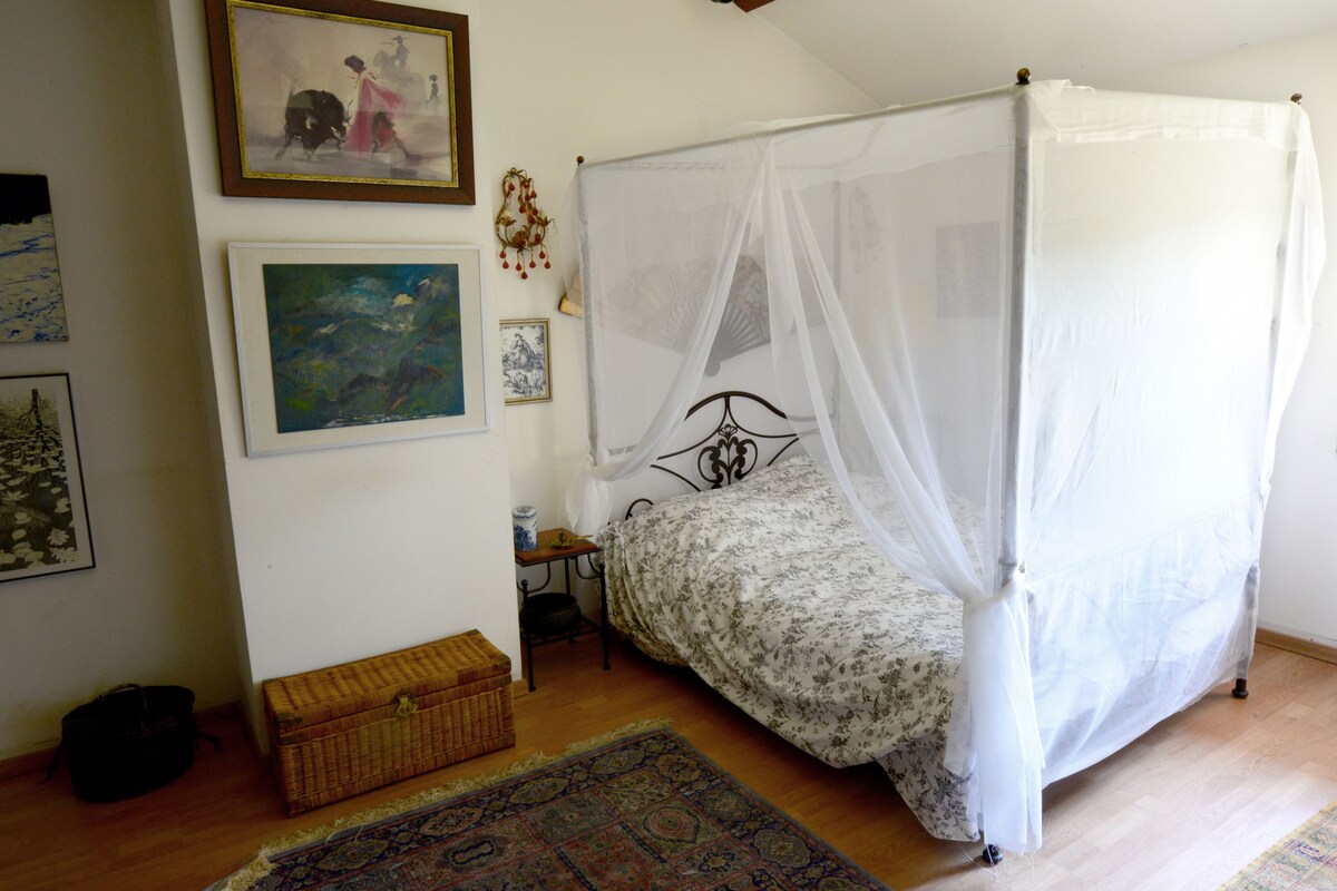 A bedroom with a four-poster canopy bed, a small bedside table, a wicker chest, and a patterned rug.