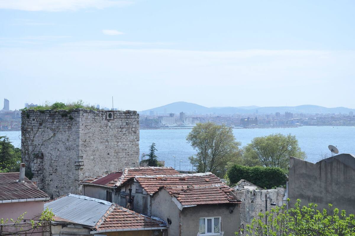 A stone tower with a tiled roof, surrounded by residential buildings with similar roofs, overlooking a body of water and a distant cityscape.