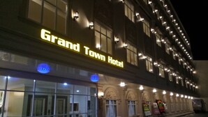 El exterior del Hotel Grand Town con señalización iluminada, ventanas amplias y una fila de mesas y sillas afuera.