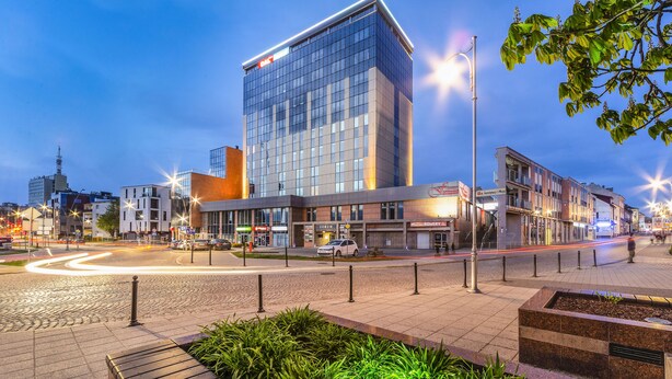 A modern building with a glass facade, located in an urban setting with a paved plaza and street lights. - Hotel Dal Kielce (Kielce)