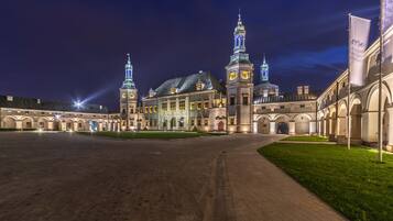 A historic building with a central dome and two towers, surrounded by a paved courtyard and a grassy area.