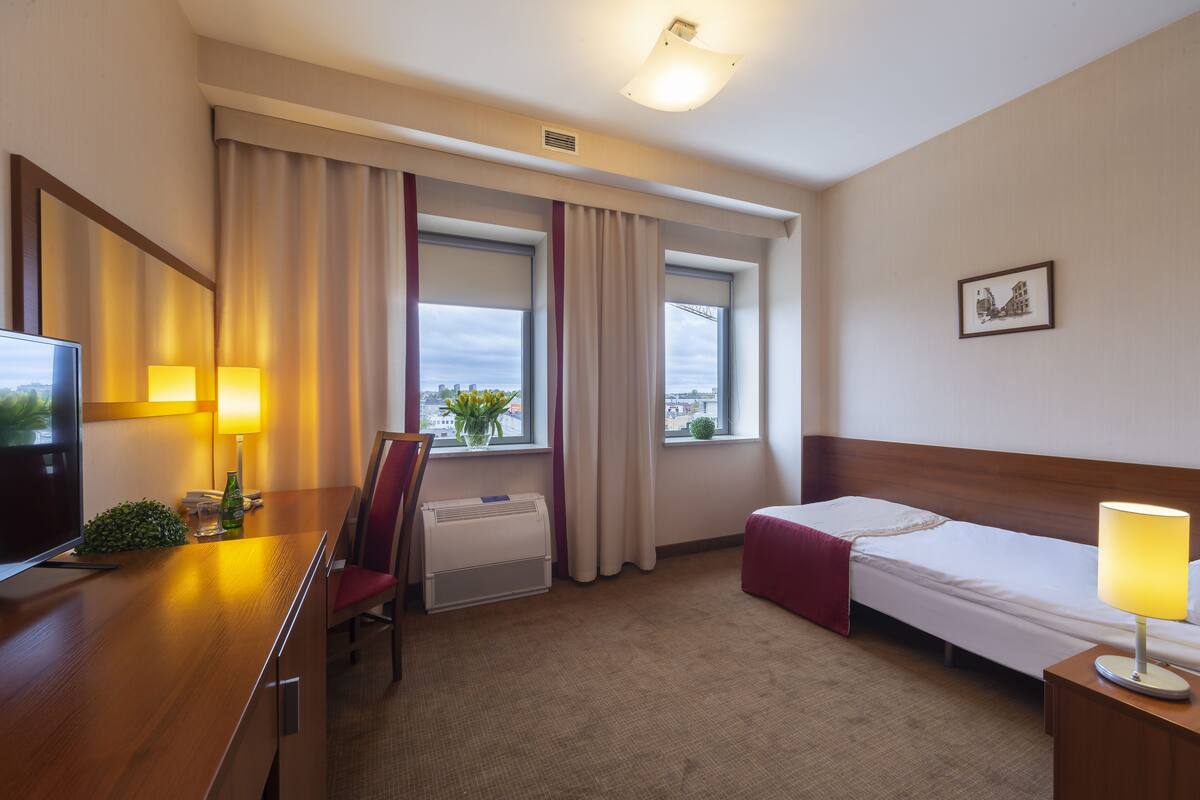 A hotel room with a bed, a desk with a chair, a TV, and a window with curtains.