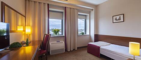A hotel room with a bed, a desk with a chair, a TV, and a window with curtains.