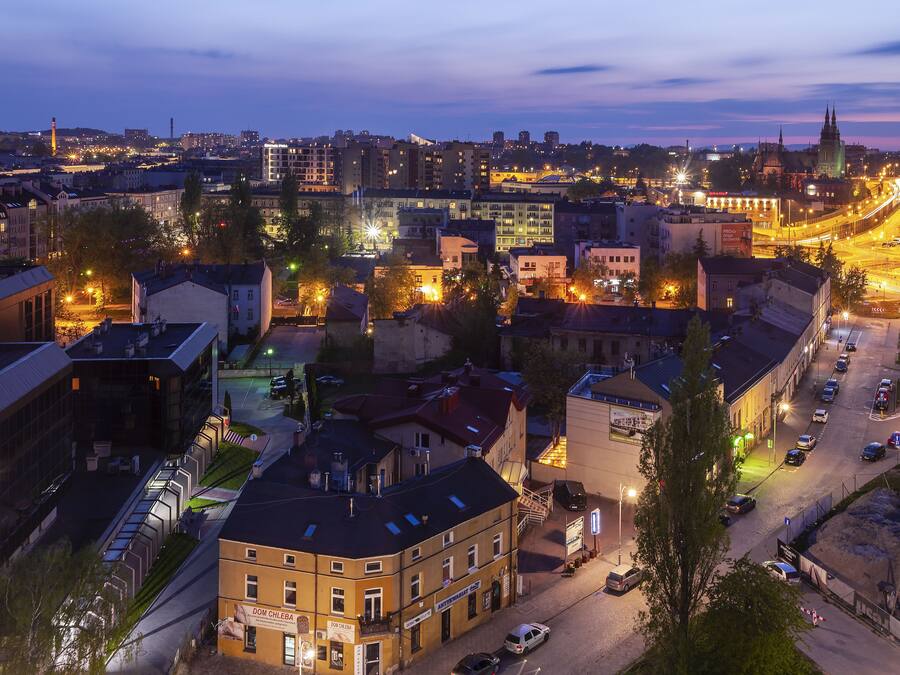 A cityscape at dusk with illuminated buildings, a prominent church spire, and a busy street.