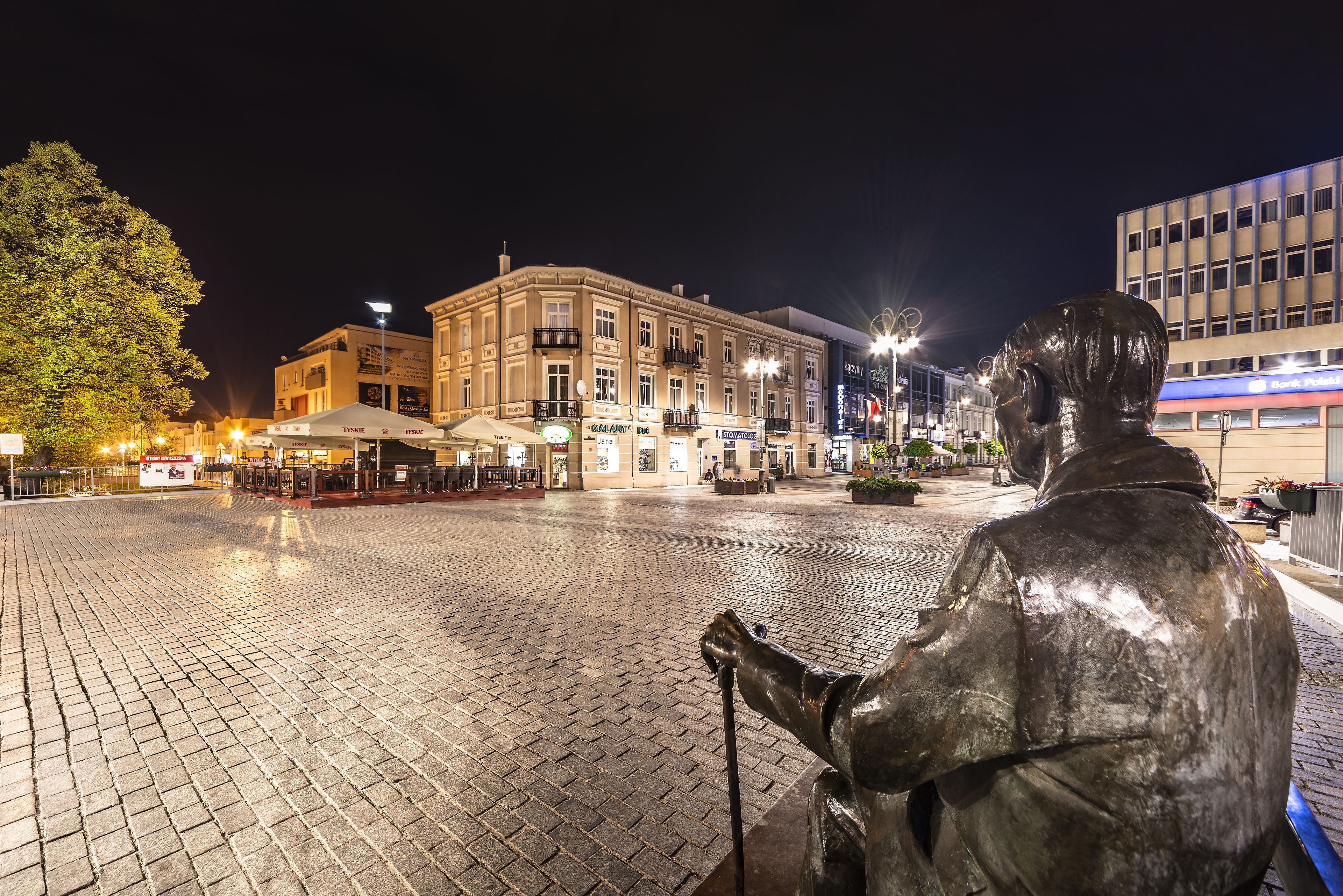 Estátua de uma figura sentada com bengala numa praça pública à noite.