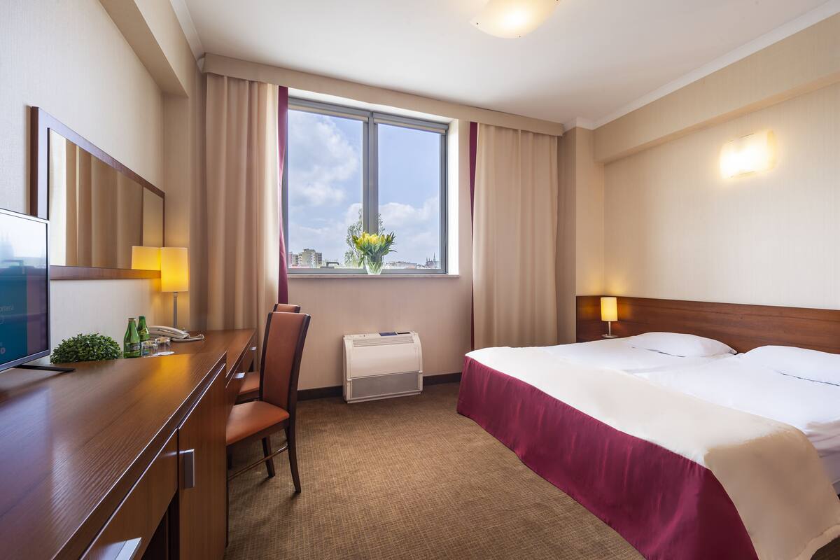 A hotel room with a bed, a desk with a chair, a TV, a window with curtains, and a wall-mounted lamp.