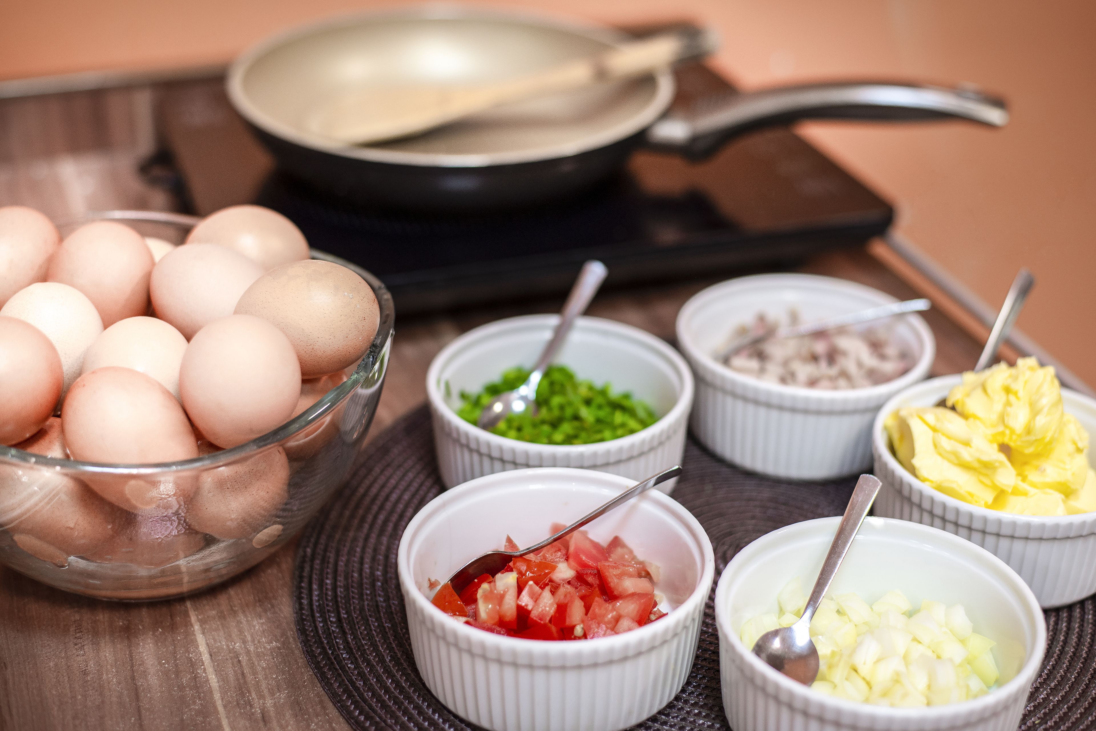 A kitchen counter with a bowl of eggs, a frying pan, and small bowls containing chopped vegetables.