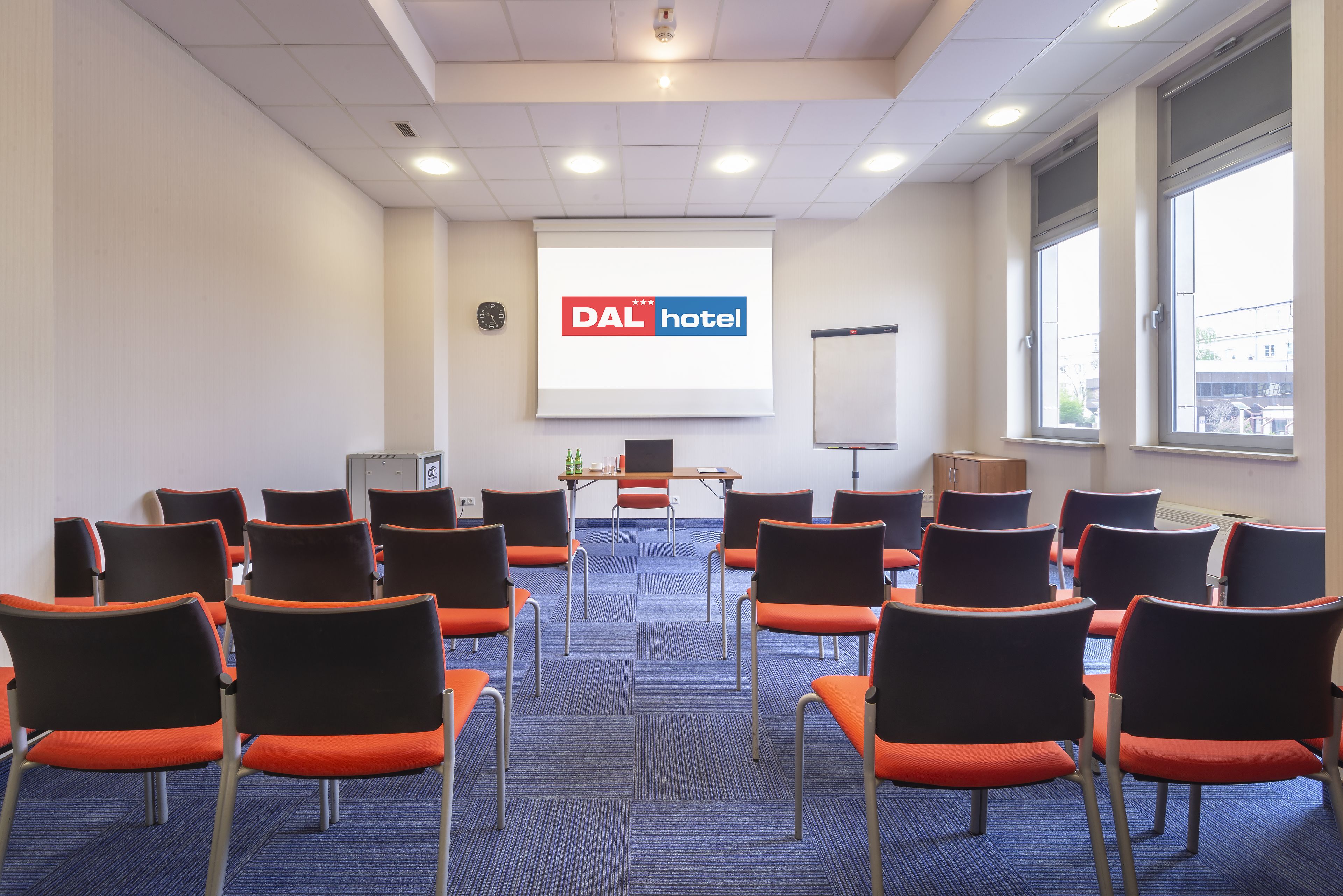 A conference room with rows of orange and black chairs, a projector screen displaying "DAL hotel", and a whiteboard.