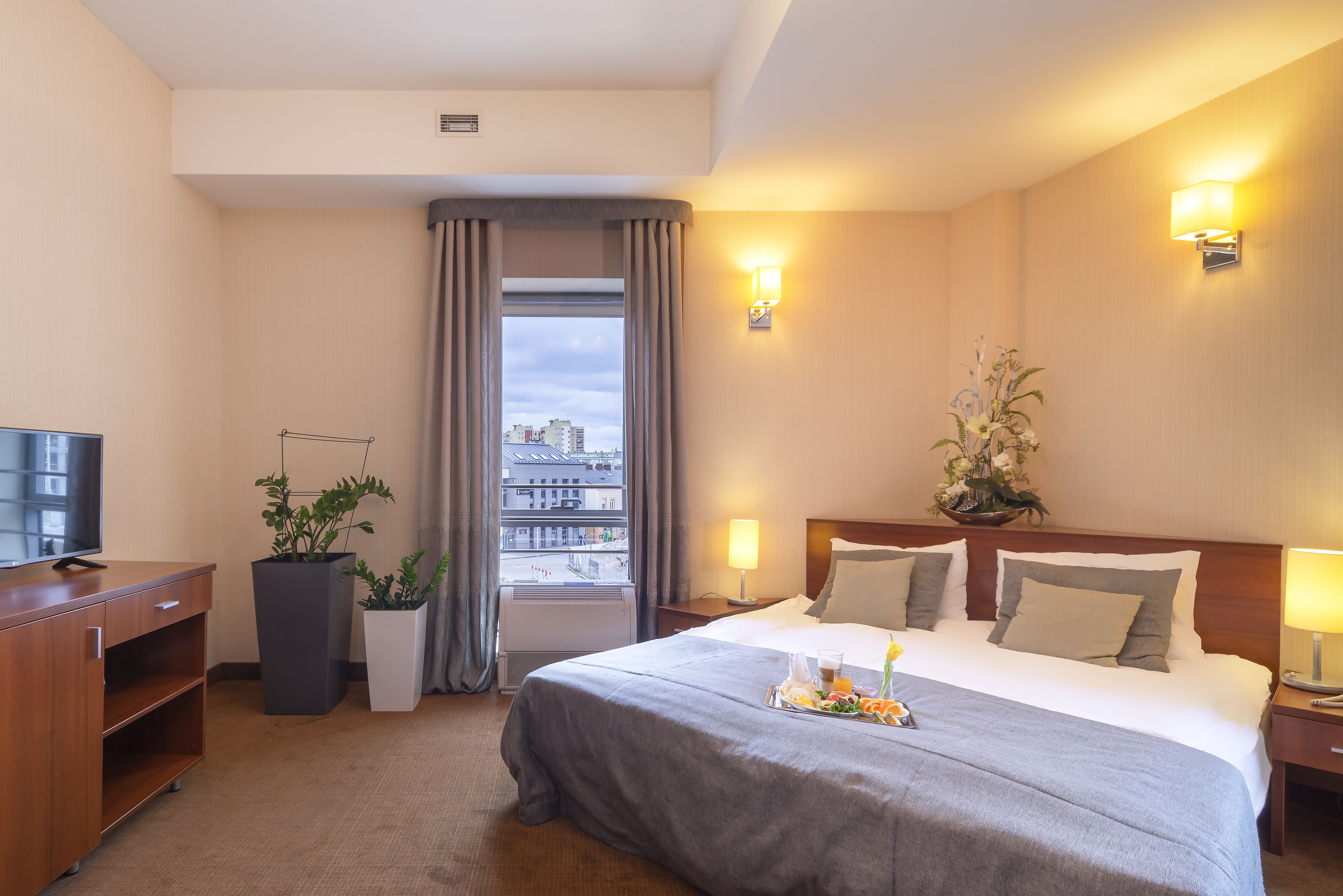 Ein Hotelzimmer mit einem großen Bett, einem Flachbildfernseher, einer Topfpflanze, einem Fenster mit Vorhängen und Blick auf die Stadt.